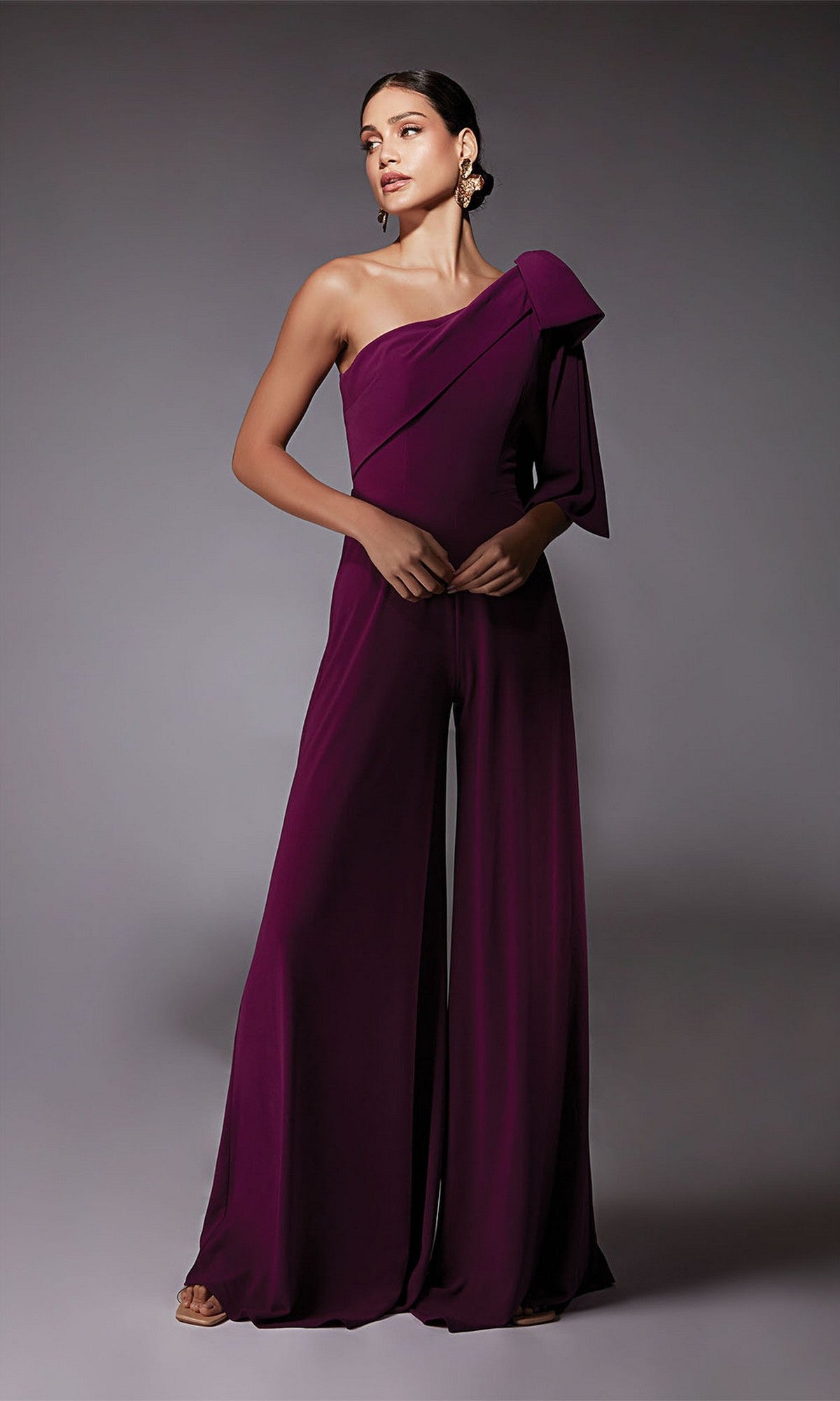 A woman wears the Alyce 70065 Long Formal Jumpsuit in Sparkling Grape—sleeveless, wide-leg, one-shoulder with a large bow—standing against a plain gray background, offering a chic alternative to a cocktail dress.